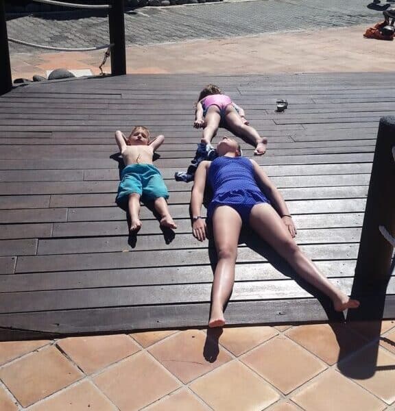 Three people, two children and one adult, lie on their backs on a sunlit wooden decking with their arms and legs relaxed, appearing to sunbathe. The scene is outdoors near tiled ground, with shadows and buildings in the background.