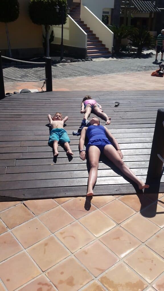 Three people, two children and one adult, are lying on their backs on a wooden decking in sunlight. The adult is wearing a blue swimming costume, and the children are in swimwear. Shadows indicate a bright, sunny day. Buildings and trees are in the background.