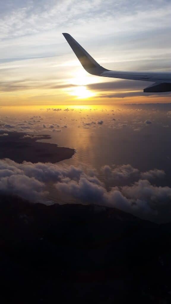 View from an aeroplane window showing a wing above clouds, with a scenic sunset over the ocean and coastline below. The sun is low on the horizon, reflecting golden light on the water, while clouds and landforms are partly visible.