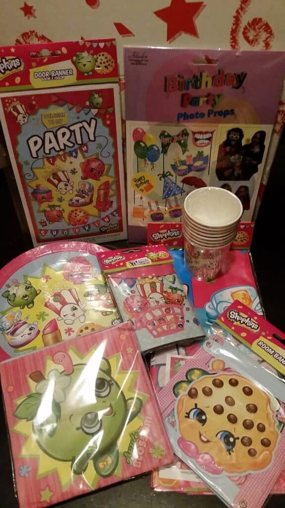 A colourful assortment of Shopkins-themed party supplies, including plates, napkins, cups, party hats, a door banner, birthday photo props, and goodie bags, all featuring cartoon food characters, arranged on a dark surface with a red and white background.