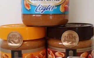 Three jars of Calvé peanut butter are stacked. The top jar has a blue lid and label marked "Light 30% less fat." The bottom left jar has an orange lid and label. The bottom right jar has a brown lid and label stating "Smooth." Peanuts are shown on the labels.