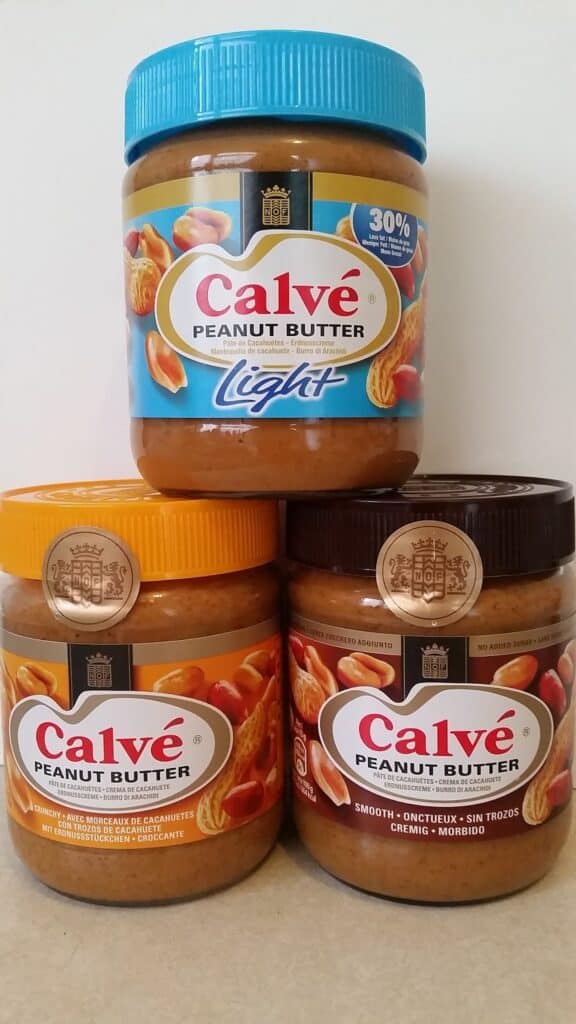 Three jars of Calvé Peanut Butter are stacked in a pyramid. The top jar is labelled "Light" with a blue lid, while the bottom two have orange and brown lids, labelled as regular and "Smooth" varieties. All feature images of peanuts and similar branding.