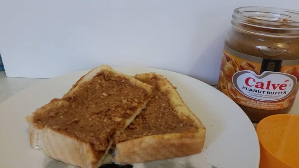 Two slices of white bread spread with crunchy peanut butter are on a white plate. To the right, there is an open jar of Calvé peanut butter with its yellow lid beside it, all against a plain light background.