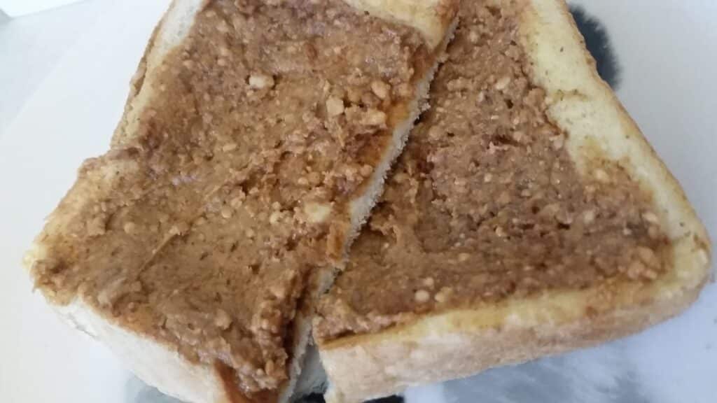 A close-up of a slice of white bread topped with crunchy peanut butter, cut diagonally in half, sitting on a white plate. The peanut butter appears thick with visible peanut pieces, covering most of the bread’s surface.