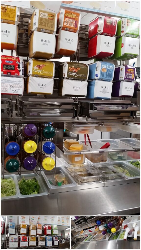A fast food prep station with various coloured condiment boxes above metal trays filled with lettuce, tomatoes, cheese, gherkins, and meats. Sauce dispensers hang on the side, and menus are visible at the top edge of the image.