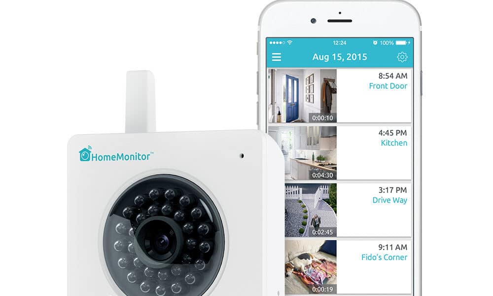 A white HomeMonitor security camera sits beside a smartphone displaying its surveillance app with four video feeds labelled Front Door, Kitchen, Driveway, and Fido’s Corner. Text above reads “No Monthly Fee,” with icons of a camera and calendar showing 7 days.