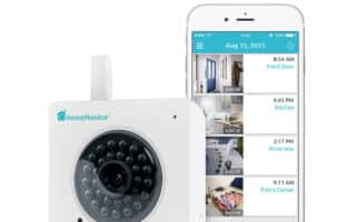 A white HomeMonitor security camera sits beside a smartphone displaying its surveillance app with four video feeds labelled Front Door, Kitchen, Driveway, and Fido’s Corner. Text above reads “No Monthly Fee,” with icons of a camera and calendar showing 7 days.