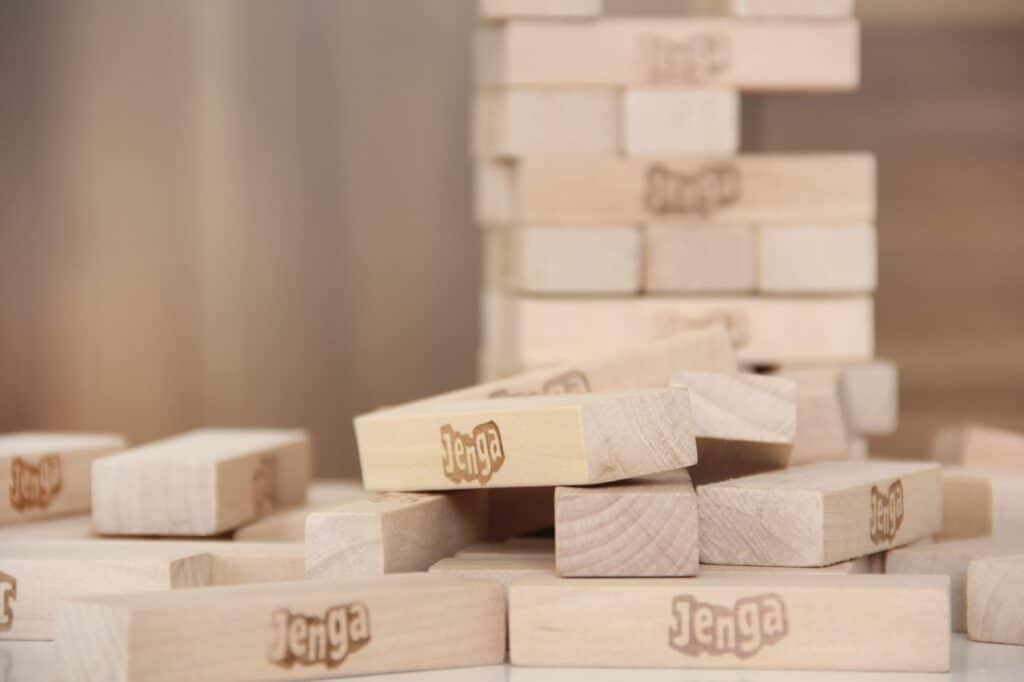 A close-up of scattered wooden Jenga blocks with several stacked and others lying in a heap. Each block is light-coloured wood with the "Jenga" logo printed on it. In the background, a partially built Jenga tower is visible.