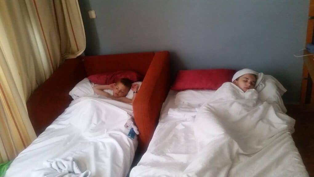 Two children lie in separate beds placed side by side in a room. Both are covered with white duvets. The child on the left is awake and looking forward; the child on the right sleeps with a towel wrapped around their head. Light enters through beige curtains.