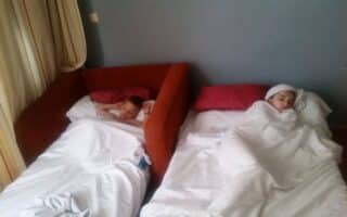Two children lie in separate beds placed side by side in a room. Both are covered with white duvets. The child on the left is awake and looking forward; the child on the right sleeps with a towel wrapped around their head. Light enters through beige curtains.