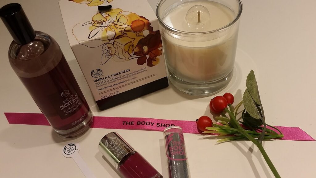 A display of The Body Shop products, including a Vanilla & Tonka Bean scented candle, a fragrance mist, lipstick, nail varnish, a pink ribbon, a faux branch with red berries and green leaves, and a cardboard box with floral artwork on a white surface.