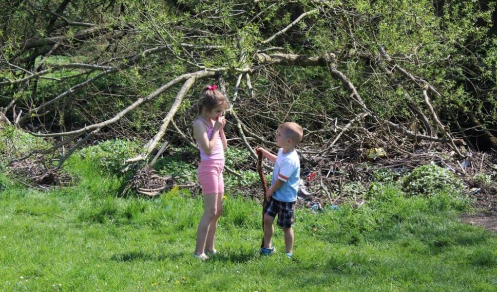 A young girl and a young boy stand on green grass in front of leafy trees and branches. The girl wears a pink outfit and holds her hand to her mouth. The boy, in a T-shirt and shorts, holds a stick and looks up at the girl. It is sunny outside.