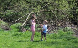 A young girl and a young boy stand on green grass in front of leafy trees and branches. The girl wears a pink outfit and holds her hand to her mouth. The boy, in a T-shirt and shorts, holds a stick and looks up at the girl. It is sunny outside.