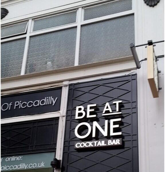 Shopfront of "Be At One Cocktail Bar" with a black sign and white letters above the entrance, plus a red neon sign below reading "OPEN TIL LATE." The building has large windows and decorative moulding along the top.