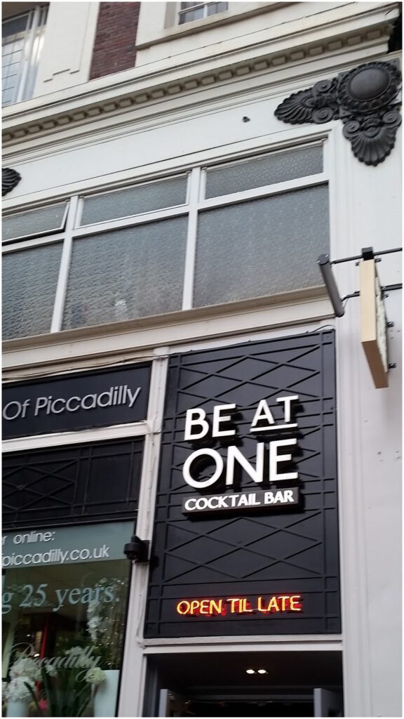 A sign reading “BE AT ONE COCKTAIL BAR” is mounted on a textured black wall above a doorway. Below it, a small neon sign says “OPEN TILL LATE.” The building has large windows and ornate black decorations above the signs.