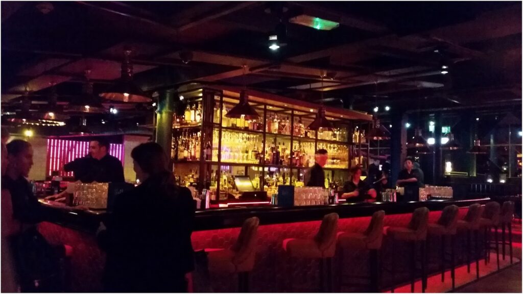 A dimly lit bar with shelves of liquor bottles behind the counter, illuminated by warm yellow and red lights. Bartenders serve drinks to customers seated on cushioned barstools. The atmosphere is lively and modern, with a few people chatting at the bar.