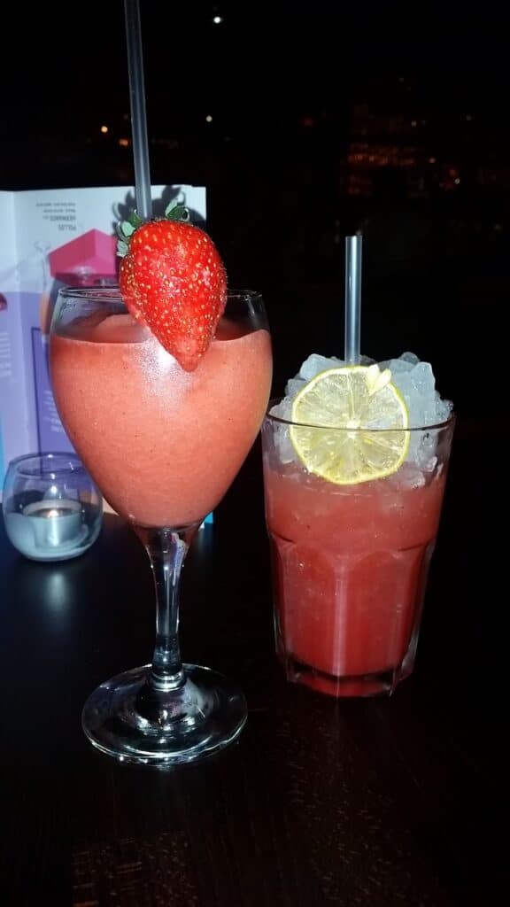 Two red cocktails on a dark table: one is a frozen drink in a wine glass garnished with a whole strawberry, the other is served over ice in a short glass with two lemon slices. A candle and colourful menu are blurred in the background.
