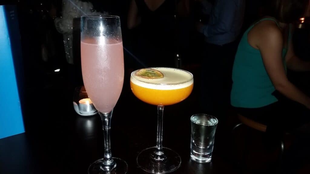 Two cocktails and a shot glass on a dark bar table: one pink drink in a champagne flute, an orange drink in a coupe glass garnished with a passion fruit slice, and a clear shot. A candle burns in the background; people sit and stand in dim lighting.