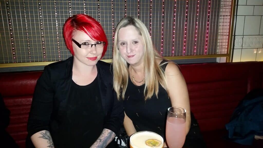 Two women sit closely on a red bench in a dimly lit bar. The woman on the left has short bright red hair, glasses, and a black outfit. The woman on the right has long blonde hair, wears a black top, and is smiling. Two pink drinks are on the table.