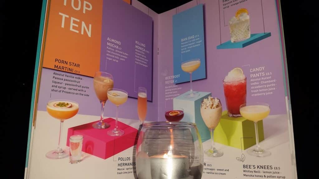 A close-up of a colourful cocktail menu stands behind a lit candle in a glass holder. The menu shows photos and descriptions of various cocktails, such as Porn Star Martini, Almond Mocha, and Bee’s Knees, with each drink displayed on vibrant geometric backgrounds.