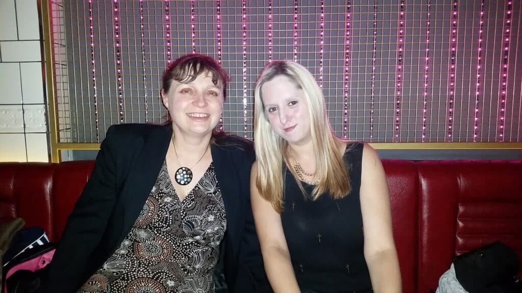 Two women sit side by side on a red bench in a dimly lit room with a wall of vertical pink lights behind them. One woman has brown hair, wears a patterned dress and black jacket; the other has long blonde hair and wears a sleeveless black dress.