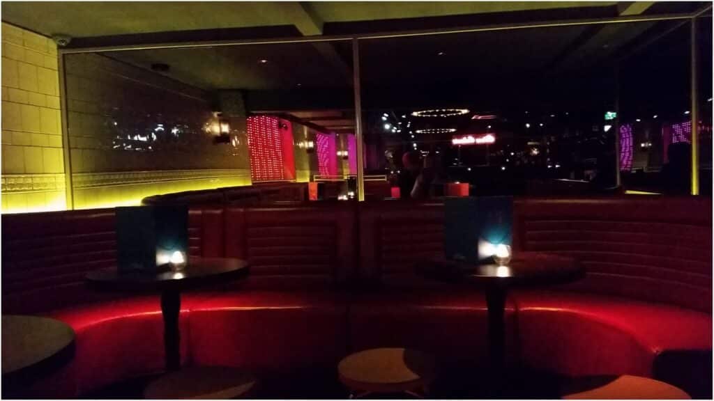 A dimly lit lounge with red cushioned booths, small round tables, and candles. Purple and yellow lighting creates a cosy ambience. The walls are lined with mirrors and tiles, reflecting the modern decor and warm hues throughout the space.