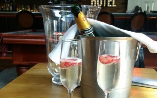 Two glasses of sparkling wine with strawberry slices sit on black serviettes on a wooden table. Behind them is a chilled bottle in an ice bucket, draped with a white cloth. The background shows a bar with the sign “Penta Hotel” and a TV showing a football match.