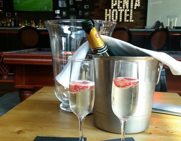 Two glasses of sparkling wine with strawberry slices sit on black serviettes on a wooden table. Behind them is a chilled bottle in an ice bucket, draped with a white cloth. The background shows a bar with the sign “Penta Hotel” and a TV showing a football match.
