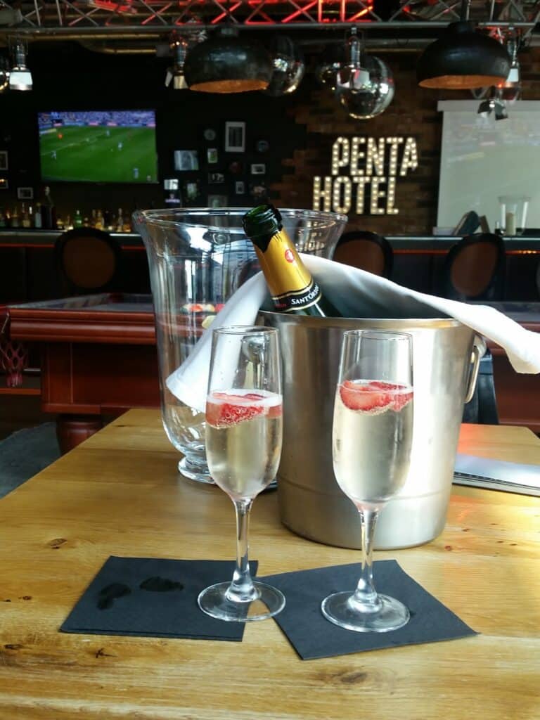A wooden table holds two champagne glasses with strawberries, a metal ice bucket with a chilled bottle of sparkling wine, and two black serviettes. In the background, a bar with "PENTA HOTEL" sign, a TV showing football, and decorative elements are visible.
