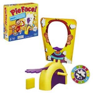 Image of the Pie Face game set, showing the colourful game box, the face cut-out frame with cartoon graphics, a purple hand for holding whipped cream, a spinner, and a yellow base. The game is designed for fun and laughter.