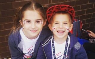Two smiling children sit on a bench in front of a brick wall. They have face paint with red and white crosses on their cheeks. The girl has long brown hair and wears a grey hoodie, while the boy wears a red cap and a blue jacket.