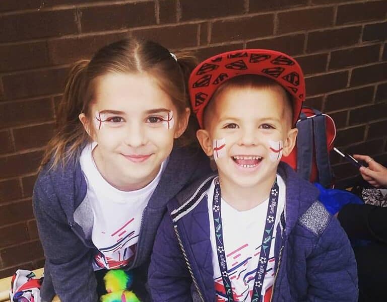 Two smiling children sit on a bench in front of a brick wall. They have face paint with red and white crosses on their cheeks. The girl has long brown hair and wears a grey hoodie, while the boy wears a red cap and a blue jacket.