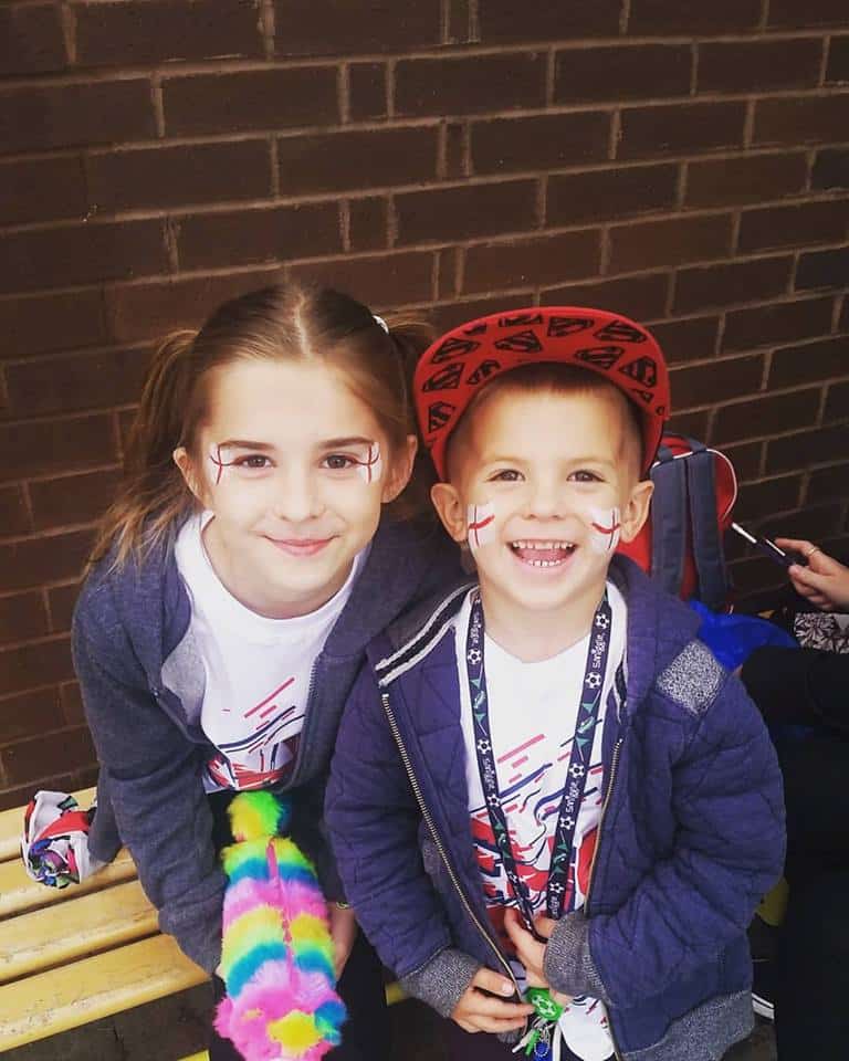 Two smiling children sit on a bench in front of a brick wall. They have face paint with red and white crosses on their cheeks. The girl has long brown hair and wears a grey hoodie, while the boy wears a red cap and a blue jacket.