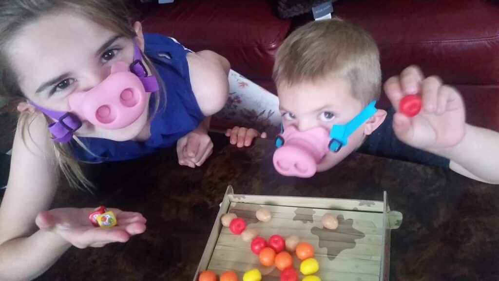 Two children wear pink pig nose masks, leaning over a table while playing a game with colourful plastic balls and a small tray. The girl holds a ball in her hand and smiles at the camera; the boy also holds a ball and looks up playfully.
