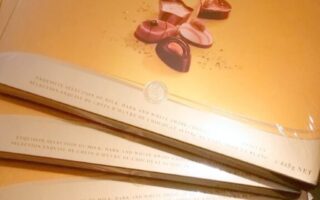 Four large, gold Lindt Swiss Luxury Selection chocolate boxes are stacked on a dark surface. The boxes feature elegant writing and images of assorted chocolates, with shiny gold accents giving a premium look.