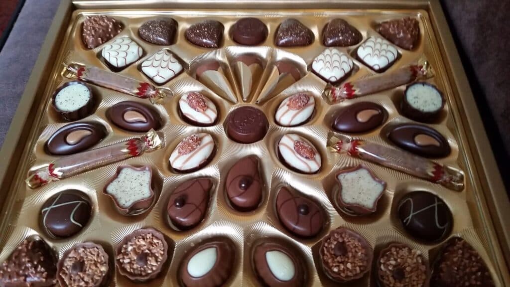A golden box filled with assorted chocolates in various shapes—hearts, ovals, stars, and rectangles. Each piece features unique decorative patterns, including swirls, lines, nuts, and white or dark chocolate accents. Two caramel-wrapped sweets are included.