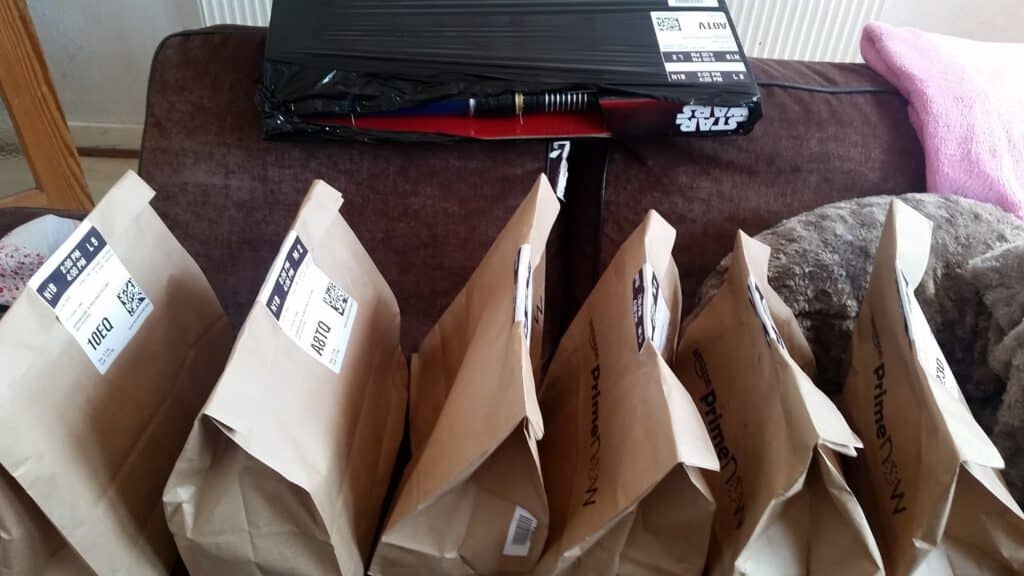 Seven brown paper delivery bags with shipping labels are lined up on a brown sofa. Behind them, there is a large rectangular parcel wrapped in black plastic, with part of a red and blue "Star Wars" lightsabre toy visible inside. A pink blanket is in the corner.