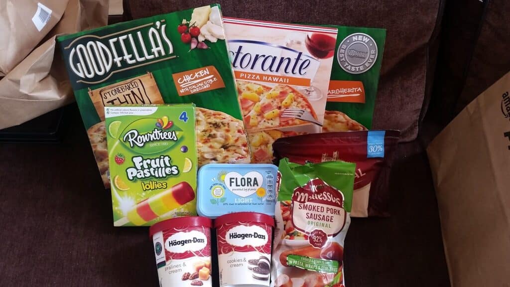A selection of groceries on a brown surface, including two frozen pizzas (Goodfella’s Chicken, Ristorante Hawaii), Rowntree’s Fruit Pastilles lollies, Flora Light spread, McCain’s smoked pork sausage, and two Häagen-Dazs ice cream tubs (Cookies & Cream, Strawberry Cheesecake).
