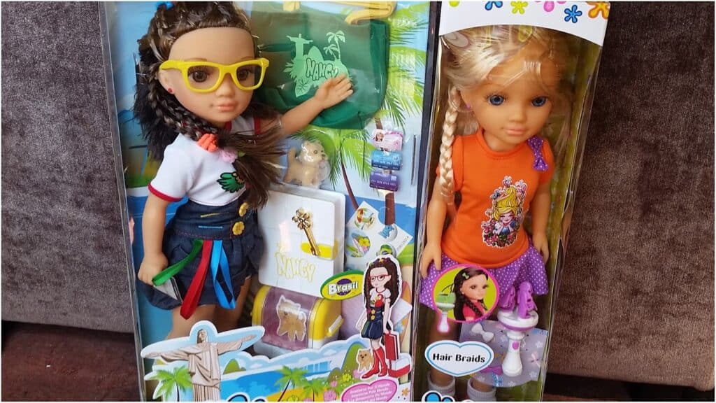 Two dolls in boxes are shown. The left doll has dark plaited hair, yellow glasses, a white shirt, blue skirt, and travel-themed accessories. The right doll has blonde hair in plaits, wears an orange top and purple skirt, and comes with a comb and hair plaiting tool.
