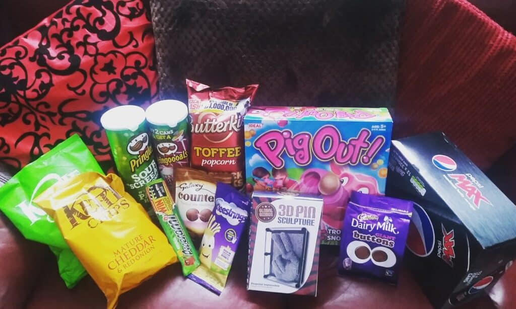 A collection of snacks and treats on a red sofa, including Pringles sour cream, Butterkist Toffee Popcorn, Metcalfe’s Cheddar popcorn, Cadbury chocolate, Rowntree’s bars, a Pig Out! game, a 3D pin sculpture toy, and a 10-pack of Pepsi Max cans.