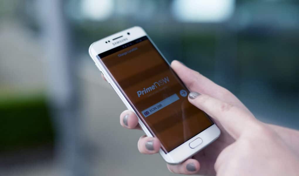 A person with grey-painted nails holds a white Samsung mobile phone, using their right index finger to interact with the Amazon Prime Now app on the screen. The background is blurred, emphasising the phone and hand.