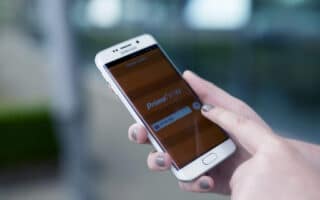 A person with grey-painted nails holds a white Samsung mobile phone, using their right index finger to interact with the Amazon Prime Now app on the screen. The background is blurred, emphasising the phone and hand.