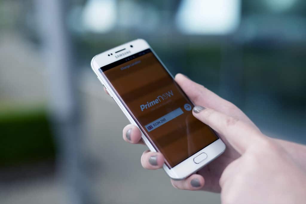A person with painted nails holds a white Samsung mobile, using their right index finger to tap the screen. The phone displays the Prime Now app with a login screen, partially blurred, outdoors with green and grey in the background.