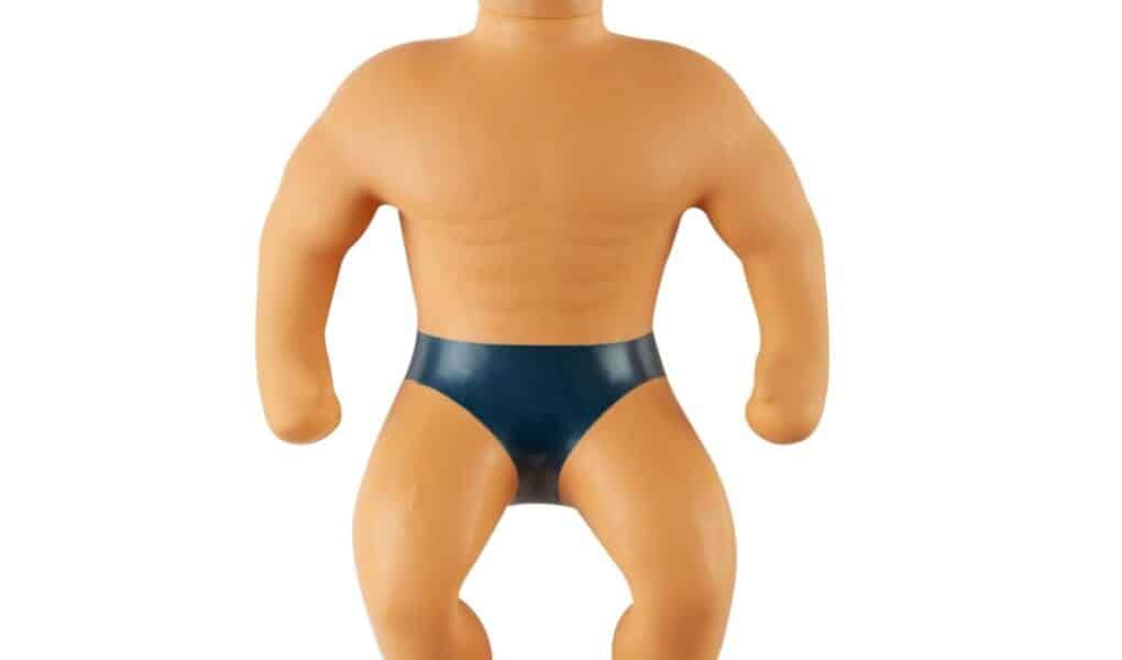 A toy figure with short blond hair, a muscular build, and a neutral expression. The figure is wearing only dark blue pants, has defined chest and arm muscles, and appears to be made of a flexible, rubbery material.