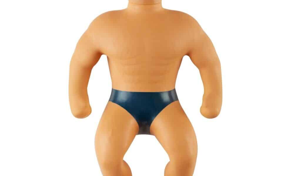 A toy figure with short blond hair, a muscular build, and a neutral expression. The figure is wearing only dark blue pants, has defined chest and arm muscles, and appears to be made of a flexible, rubbery material.