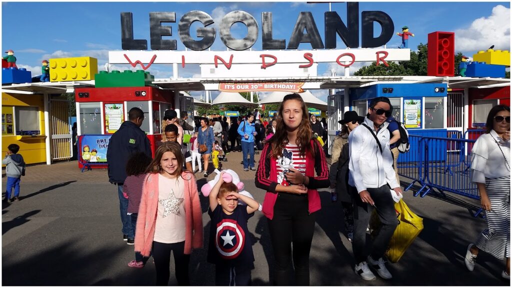 Three children stand in front of the LEGOLAND Windsor entrance, posing for a photo. The entrance sign is large and colourful, with crowds of people walking in and around the entrance under a sunny blue sky. The children appear excited and are wearing casual clothes.