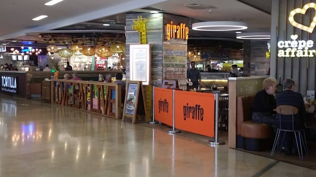A casual restaurant area in a shopping centre features a "giraffe" eatery with rustic wood decor and orange signs. People are seated at tables eating. Nearby is a "crêpeaffair" café with a heart-shaped neon sign. The floor is shiny and the space is brightly lit.
