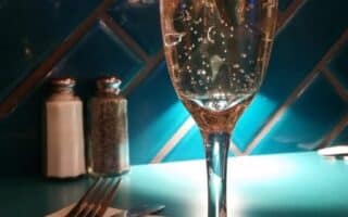 A tall champagne flute with sparkling wine sits on a teal table. Behind it are salt and pepper shakers. Fork and knife rest on a folded serviette to the left. The background features blue tiles and a wooden panel, illuminated by warm lighting.