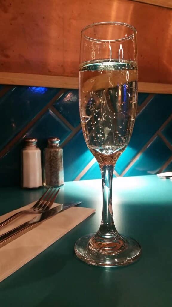 A close-up of a glass of sparkling wine on a teal table, with bubbles visible. In the background, there are salt and pepper shakers and cutlery on a white serviette. The wall has turquoise tiles in a diagonal pattern. The lighting is warm and cosy.