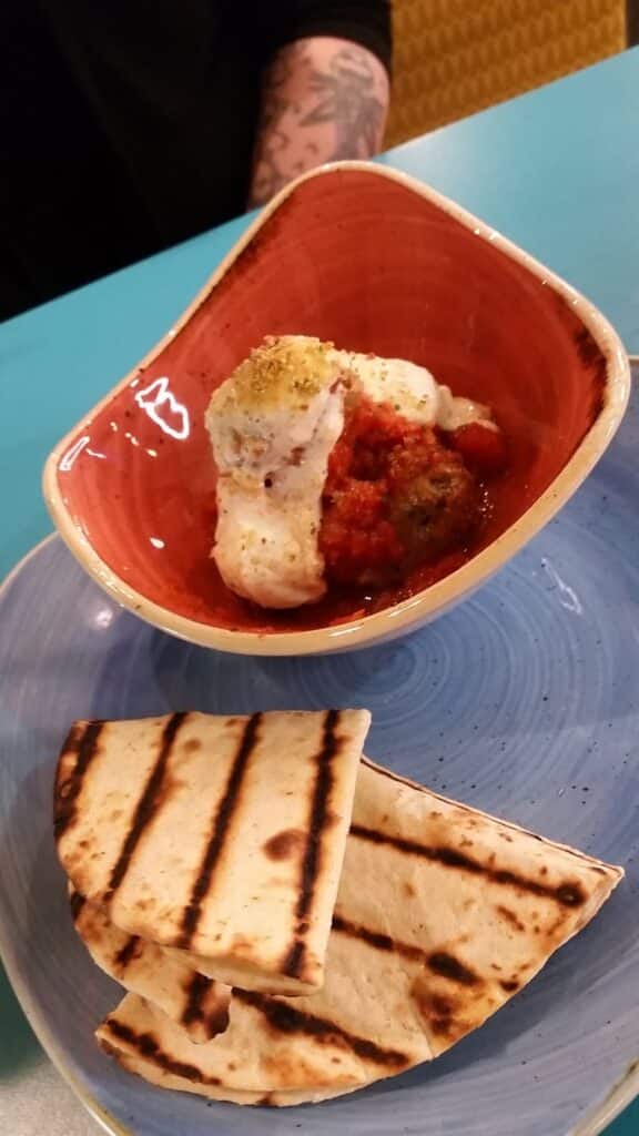 A bowl with two scoops of white cheese on red tomato sauce is placed on a blue plate, accompanied by three pieces of grilled flatbread. A person with a tattooed arm is partially visible in the background.
